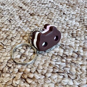 May include: A keychain featuring a dark brown, cat-shaped sandwich cookie charm with a light pink cream filling. The charm has two circular cutouts for eyes and is attached to a standard silver-toned metal split ring by a short chain link.