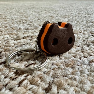 May include: A keychain featuring a dark brown, cat-shaped charm with two circular cutouts for eyes and a bright orange center layer. The charm is attached to a standard silver-colored metal split ring, providing a functional way to organize keys.
