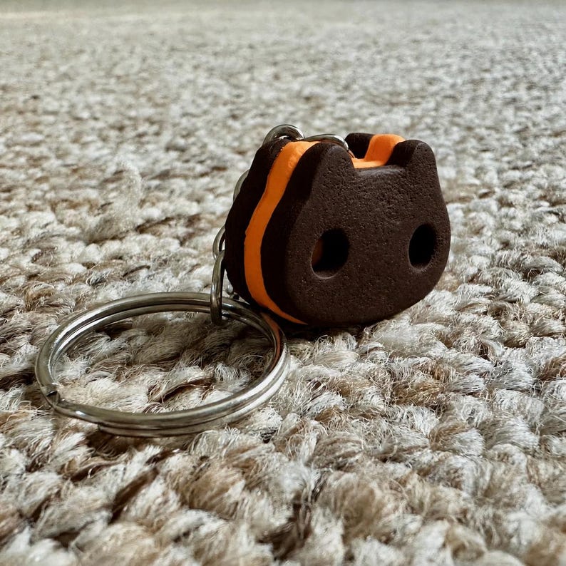 May include: A keychain featuring a dark brown, cat-shaped charm with two circular cutouts for eyes and a bright orange center layer. The charm is attached to a standard silver-colored metal split ring, providing a functional way to organize keys.