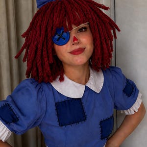 May include: A rag doll costume featuring a blue dress with white collar and cuffs, dark blue patches with black stitching, and a red triangular nose. The headpiece has red yarn hair, a blue hair bow, and a large blue button over one eye.