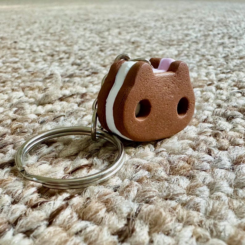 May include: A brown, cat-shaped sandwich cookie keychain featuring a white cream filling layer and two circular cutouts for eyes. The charm is attached to a standard silver-colored metal split ring for keys.