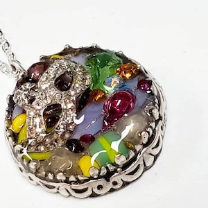 May include: A silver necklace with a round pendant featuring a silver filigree design. The pendant is filled with colorful glass pieces, including purple, green, yellow, and red.