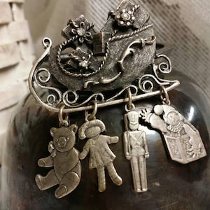 May include: Silver tone Christmas sleigh charm with dangling ornaments including a teddy bear, a doll, a toy soldier, and a jack-in-the-box.