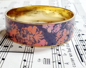 May include: A gold bangle bracelet with a floral pattern. The pattern is in shades of pink and purple on a dark background.