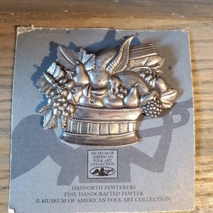 May include: A silver pewter pin depicting a basket overflowing with fruit and vegetables. The pin is a part of the Museum of American Folk Art Collection by Danforth Pewterers.