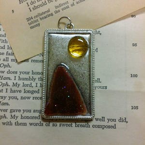 Puede incluir: Un colgante de plata con una imagen de resina marrón y dorada en su interior. La imagen representa una montaña con un sol amarillo encima. El colgante está rodeado de un marco de plata con un borde de cuentas.
