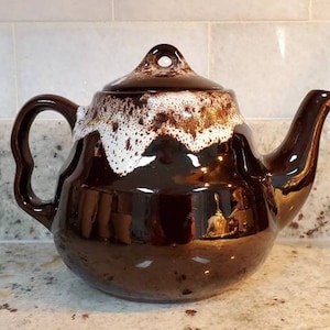 Op de afbeelding: Een bruine keramische theepot met een deksel en een gebogen handvat. De bovenkant van de theepot en het deksel hebben een wit, gestructureerd ontwerp. De theepot staat op een aanrecht.