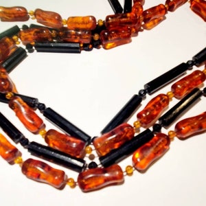 May include: A multi-strand necklace made of amber and black beads. The amber beads are a warm, honey colour and are shaped like small, irregular cylinders. The black beads are smooth and cylindrical. The necklace is a classic and elegant design.