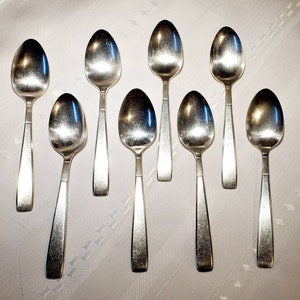 May include: Eight silver teaspoons with rounded bowls and long, slender handles. The spoons are arranged in a row on a white surface.