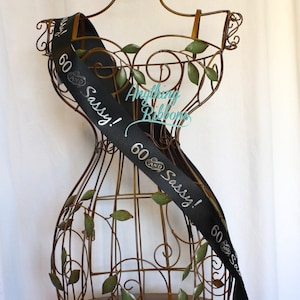 May include: Black satin sash with silver lettering that reads "60 and Sassy!"  The sash is wrapped around a decorative metal mannequin.