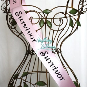 May include: Pink satin sash with the word "Survivor" printed in black on it. The sash is draped over a decorative metal stand.