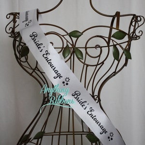 May include: White satin sash with black text that reads "Bride's Entourage" and "Anything Ribbons". The sash is displayed on a decorative metal stand.
