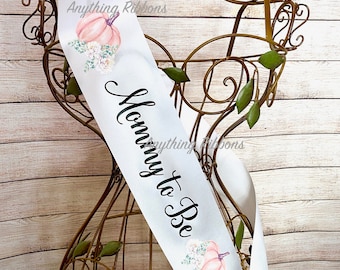 Op de afbeelding: Witte satijnen sjerp met de tekst "Mommy to Be" in zwarte letters. De sjerp is versierd met aquarel illustraties van pompoenen en bloemboeketten.