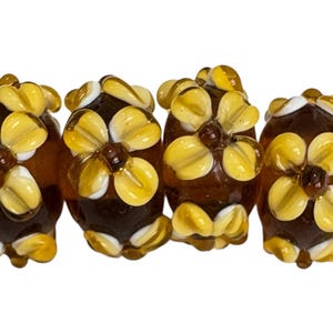 May include: Four brown glass beads with yellow and white flower designs. Each bead has a dark brown center and is strung on a red thread. The flowers have multiple petals and are arranged around the top of the beads.