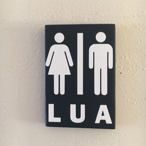 May include: A black sign with white stick figures representing a woman and a man. The sign also has the word "LUA" written in white letters.
