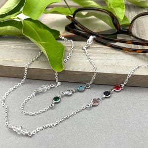 May include: A silver eyeglass chain with colourful gemstones. The chain features a series of round gemstones in various colours, including green, blue, pink, red, and black. The chain is designed to hold eyeglasses and is displayed with a pair of tortoiseshell glasses.
