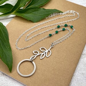 May include: A silver-colored necklace with a leaf pendant and a circular charm. The necklace features a delicate chain with small green beads. The leaf pendant is a stylized design. The necklace is displayed on a brown surface with green leaves.