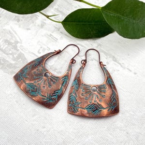 May include: A pair of copper-toned earrings featuring a geometric design with turquoise detailing. The earrings have a distinctive shape with a central hole and are decorated with leaf-like patterns. They are presented on a white surface with green leaves in the background.