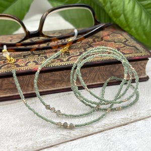 May include: A green and gold beaded eyeglass chain with a gold clasp. The chain is made of small beads and has a few larger, clear beads.