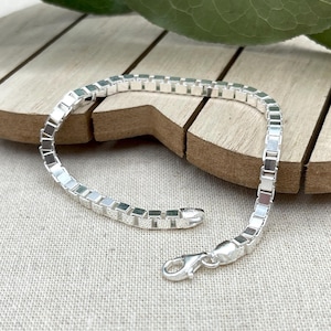 May include: A silver box chain bracelet. The bracelet is made of small, square links. It has a lobster clasp. The bracelet is displayed on a wooden surface. The bracelet's design is simple and elegant.