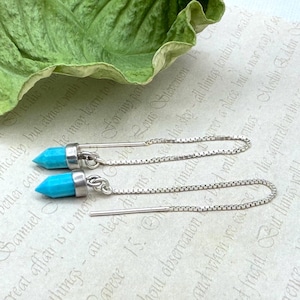 May include: A pair of turquoise and silver earrings. Each earring has a bright blue, pointed gemstone set in a silver cap, connected to a silver chain and a long, thin post. The earrings are displayed on a textured, off-white surface.