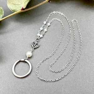 May include: A silver chain necklace with a silver heart charm and a single pearl. The chain is attached to a silver ring.
