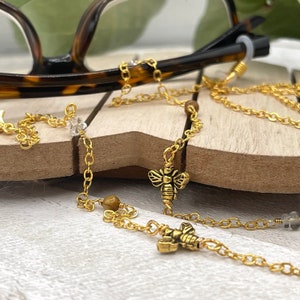 May include: Gold eyeglass chain with bee charms and tiger's eye beads. The chain is attached to the arms of a pair of spectacles.