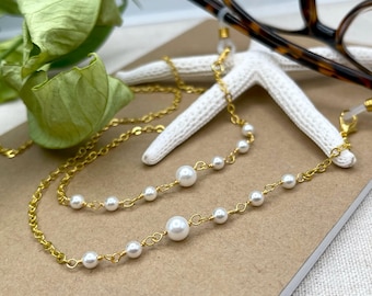 May include: A gold chain with white pearl beads, designed to hold glasses. The chain is laying on a brown surface with a white starfish.