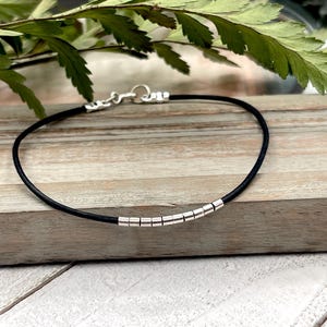 May include: A black leather bracelet with silver beads. The bracelet is on a wooden surface.