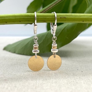 May include: Dangle earrings featuring gold-coloured discs and silver-coloured accents. The earrings have a lever-back closure and are suspended from small beads. A green stem and leaf form the background.
