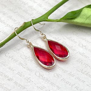 May include: Gold-coloured drop earrings featuring faceted, teardrop-shaped red gemstones. The earrings have a simple, elegant design, with a gold frame around the red stone. The earrings are displayed on a light-coloured background.