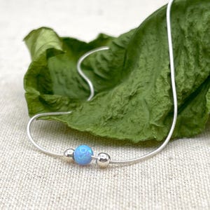 May include: A silver necklace featuring a light blue opal bead and two silver beads. The necklace is presented on a green leaf, set against a neutral backdrop. A simple and elegant piece of jewellery.