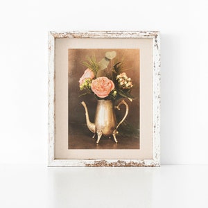 May include: A framed print of a still life featuring a silver teapot filled with pink roses and greenery. The background is a muted brown color.