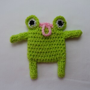 May include: A green crocheted frog toy with pink accents. The frog has large eyes and a small mouth. It has two arms and two legs.
