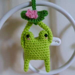 May include: A green crocheted frog holding a pink and green crocheted lily pad. The frog has black eyes and a pink mouth.