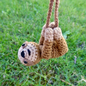 May include: A crocheted sloth toy hanging from a brown string. The sloth is brown and beige with black eyes.