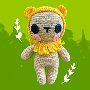 Könnte beinhalten: Gehäkelte Löwenpuppe mit gelber Kapuze und Mähne, schwarzen Augen und rosa Wangen. Das handgefertigte Spielzeug ist beige und gelb, mit einer T-förmigen Nase. Der Hintergrund ist ein grüner Farbverlauf.