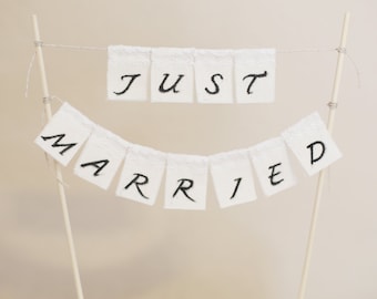 May include: White fabric banner with the words "Just Married" in black cursive lettering. The banner is strung between two wooden dowels.