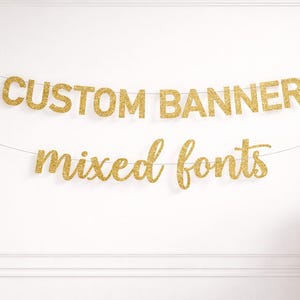 Può includere: Banner dorato glitterato con le parole "CUSTOM BANNER" in lettere maiuscole sopra "mixed fonts" in corsivo. Lo striscione è appeso a un filo d'oro sottile contro un muro bianco.