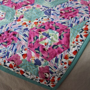 May include: A colorful quilt with a floral pattern in shades of pink, blue, and white. The quilt features a geometric design with hexagonal shapes and a teal border. The fabric has a mix of floral and abstract patterns.
