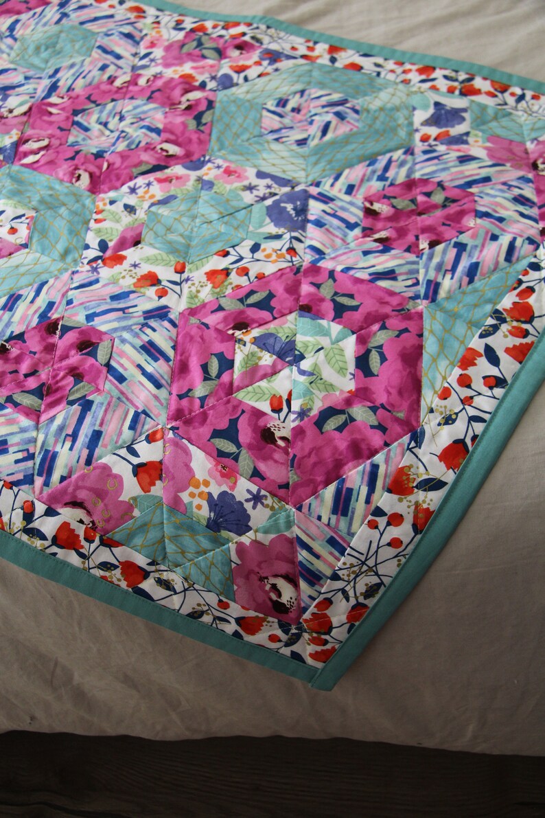 May include: A colorful quilt with a floral pattern in shades of pink, blue, and white. The quilt features a geometric design with hexagonal shapes and a teal border. The fabric has a mix of floral and abstract patterns.