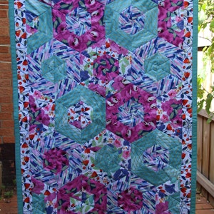May include: A colorful quilt with a floral and geometric design. The quilt features large, hexagonal shapes in teal, surrounding vibrant magenta floral patterns. The edges are trimmed with a teal border and a floral print.