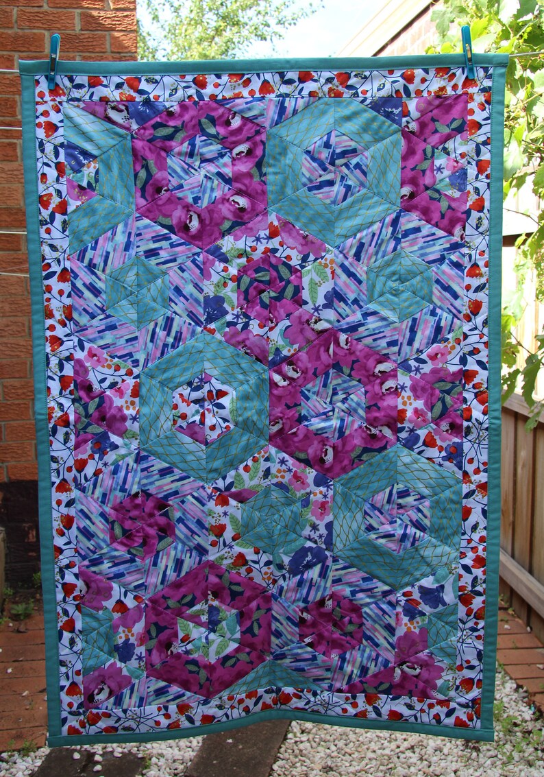 May include: A colorful quilt with a floral and geometric design. The quilt features large, hexagonal shapes in teal, surrounding vibrant magenta floral patterns. The edges are trimmed with a teal border and a floral print.