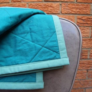 May include: A teal and light green quilted blanket draped over a brown chair. The blanket has a diamond pattern and a light green border. The background is a red brick wall.