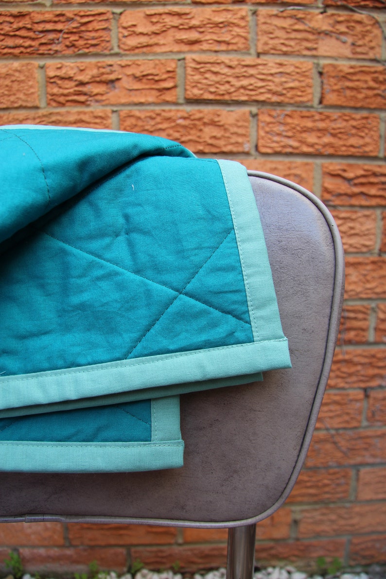 May include: A teal and light green quilted blanket draped over a brown chair. The blanket has a diamond pattern and a light green border. The background is a red brick wall.