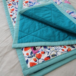 May include: A folded quilt with a floral pattern in pink, red, and blue on a white background. The quilt has a teal backing and teal binding. The quilt is made of soft fabric and is likely for a baby or child.