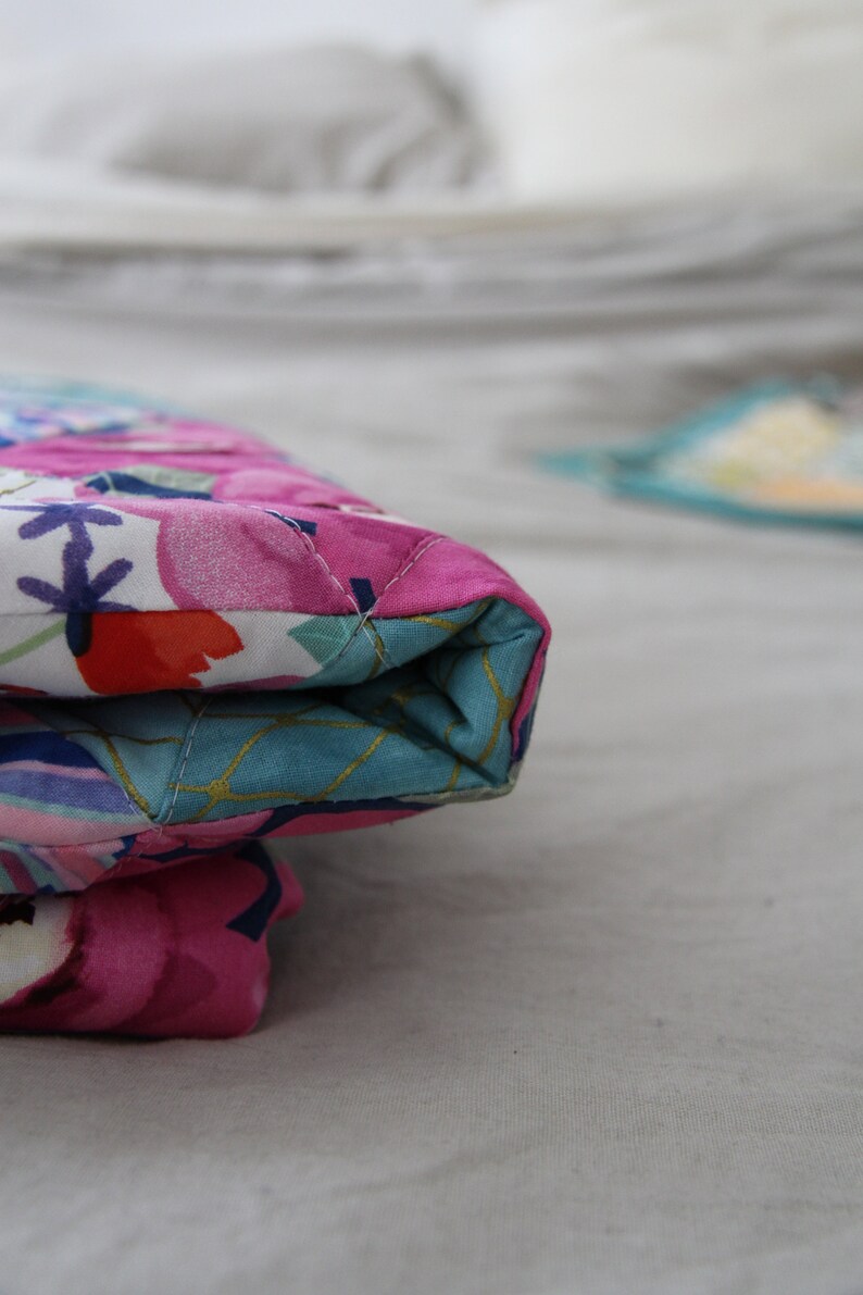 May include: Close-up of a quilted fabric item with a floral pattern in pink, white, and blue. The item is folded, revealing a turquoise interior with gold accents. The stitching is visible.