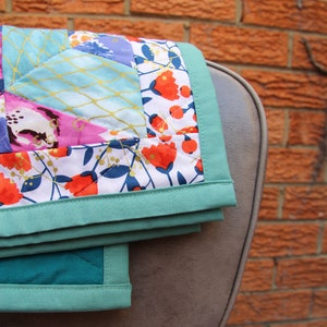May include: A folded quilt with a floral pattern in shades of red, pink, and blue, accented with gold stitching. The quilt has a teal border and is draped over a gray chair, set against a brick wall.