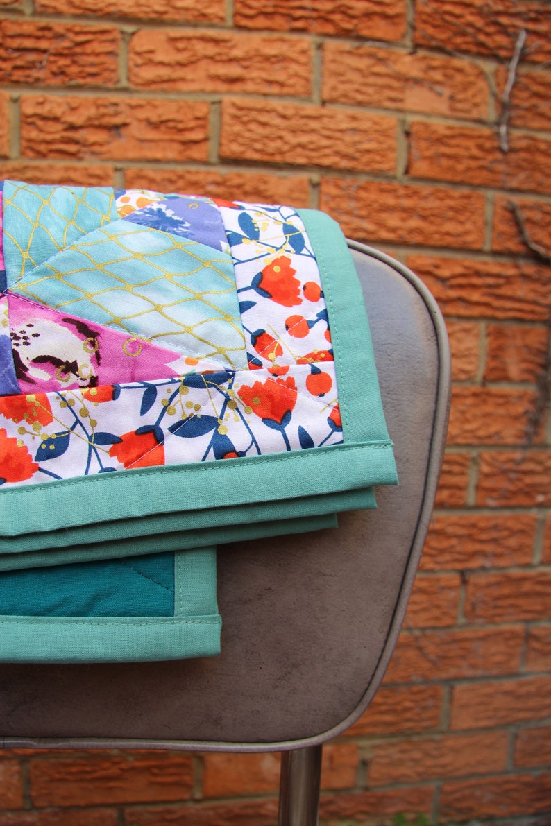 May include: A folded quilt with a floral pattern in shades of red, pink, and blue, accented with gold stitching. The quilt has a teal border and is draped over a gray chair, set against a brick wall.