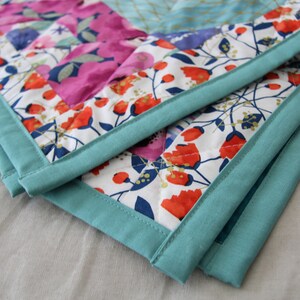 May include: Close-up of a quilted blanket with a floral pattern in pink, red, and blue on a white background. The blanket has a teal border. The quilt is folded over, showing the corner.
